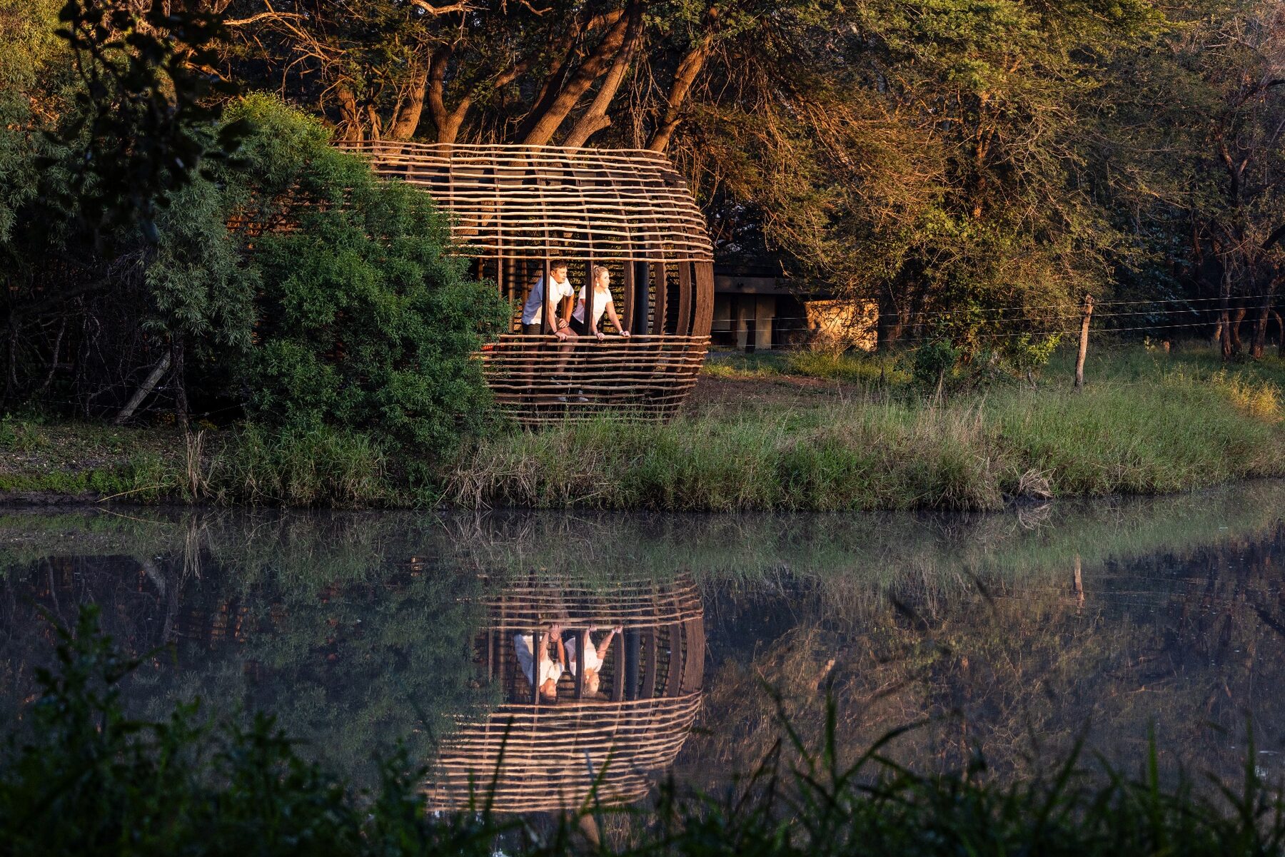 Two lodge professionals standing in a woven riverside structure reflected in the water